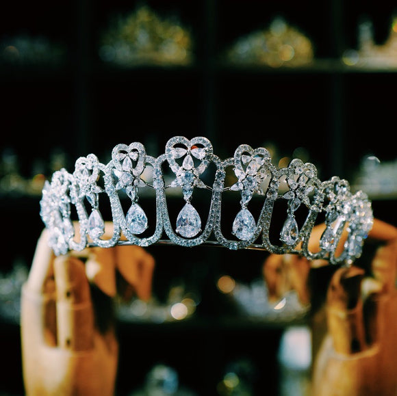  sparkling silver tiara featuring intricate heart-shaped designs and dangling teardrop zirconia stones. The large zirconia stones are suspended to create a dynamic, shimmering effect.