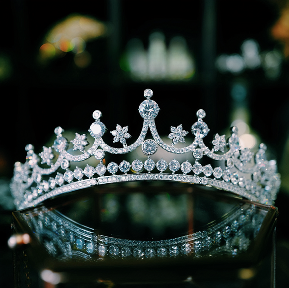 A sparkling natural zircon tiara with intricate floral and star-shaped designs, featuring brilliant, diamond-like stones set in a delicate silver frame. The crown rests elegantly on a reflective surface, showcasing its luxurious craftsmanship.